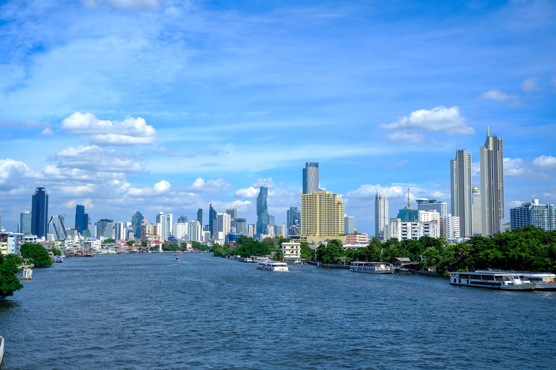 Bangkok skyline overlooking the Chao Phraya River with modern buildings and green waterfront scenery.