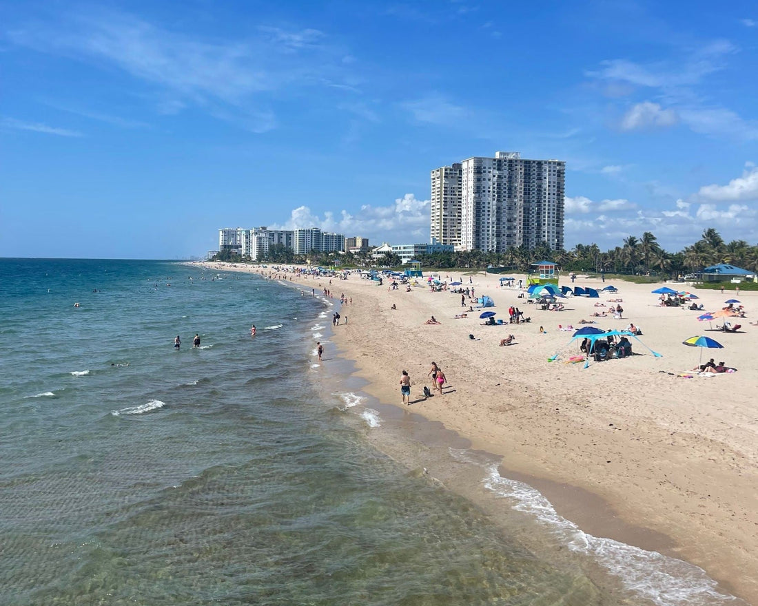 Sunny Fort Lauderdale beach with umbrellas, Atlantic Ocean water and South Florida high-rise buildings along the shoreline.