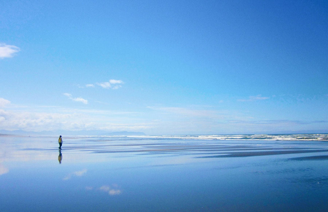 Person walking across the wide wet beach in Seaside, Oregon, with the Pacific Ocean and distant coast reflected in the sand.