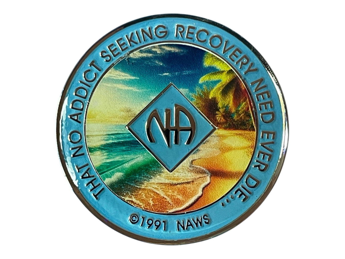 Narcotics Anonymous recovery medallion with light blue border, NA diamond logo, and tropical beach scene featuring ocean waves, palm trees, and sunset, with the phrase ‘That No Addict Seeking Recovery Need Ever Die.’