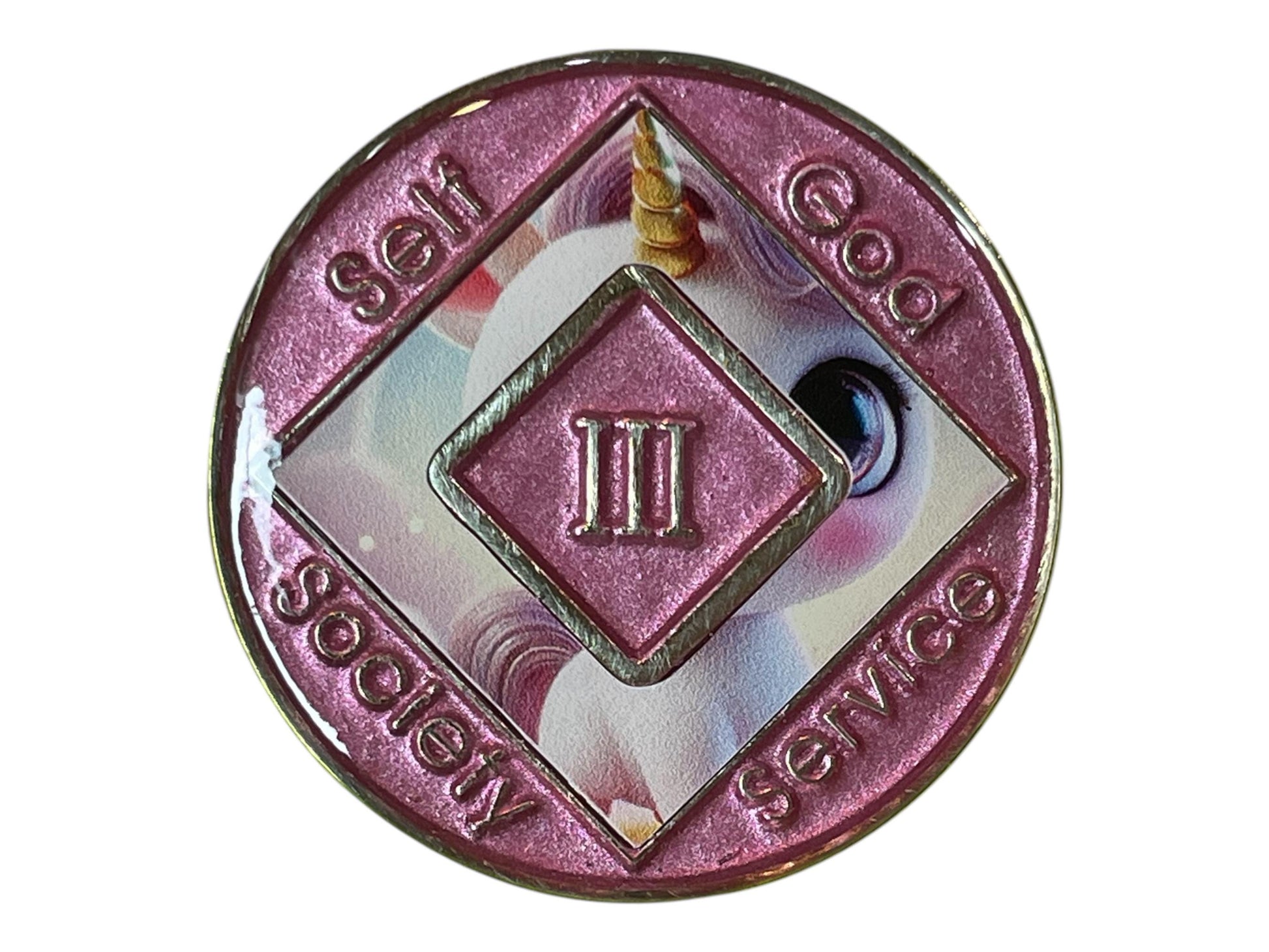 A pink metal coin with a unicorn image and a diamond shape in the center. The diamond shape has the Roman numeral 'III' in the center. The coin has the words 'self', 'society', 'service', and 'god' around the outside.