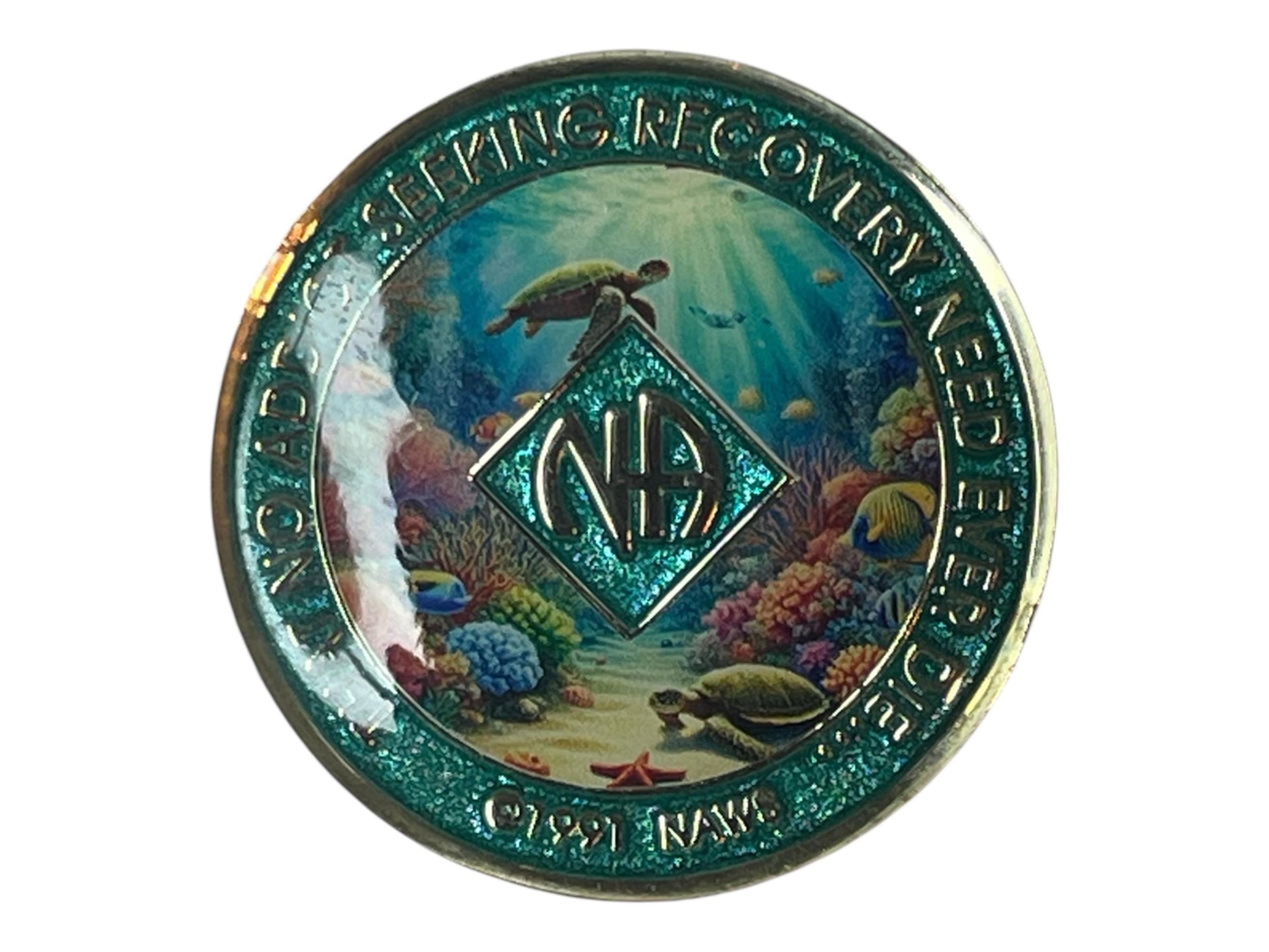 A teal-colored coin with an underwater scene featuring turtles, coral, and fish. The coin has the words 'Seeking Recovery' and 'Need Ever Die' around the edge. In the center is a teal diamond shape with the letters 'NA' inside. The bottom of the coin reads '©1991 NAWS'.